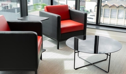 Espace café avec fauteil noir et coussin rouge. Table basse ronde devant celles-ci. Derrière, les murs sont vitrés, modernes.